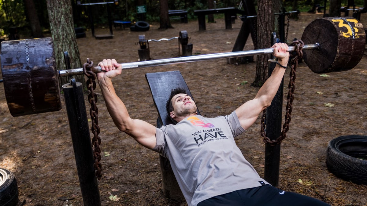 Man weightlifting outdoors, using a bench press setup amid trees for strength training.